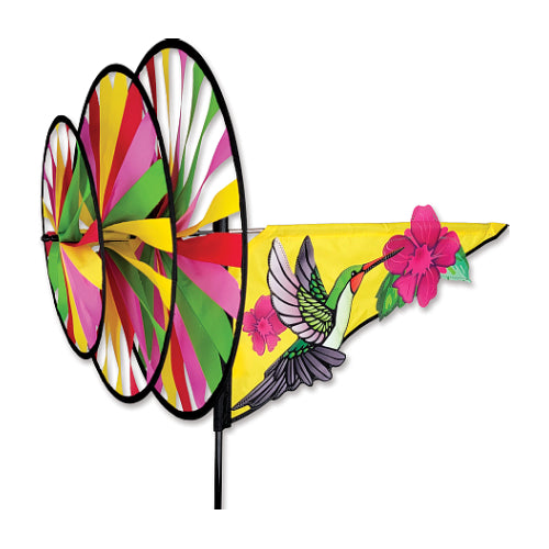 A colorful garden spinner with three large circular blades in pink, yellow, green, and white, attached to a yellow triangular pennant flag featuring a hummingbird and pink flowers, mounted on a black stake against a white background.