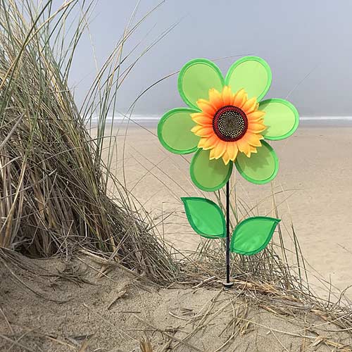 A decorative flower-shaped garden stake with green outer petals, a yellow and orange center, and two green leaves, planted in sand next to beach grass with a beach in the background.