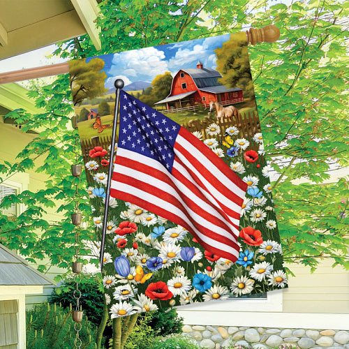 Decorative flag with American flag and scenic landscape featuring a barn and flowers, hanging outdoors.
