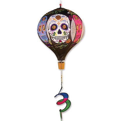 A hot air balloon-shaped hanging wind spinner decorated with colorful Dia de los Muertos sugar skull patterns, featuring a brown balloon body, a small gold basket, and a multi-colored spiral ribbon tail.