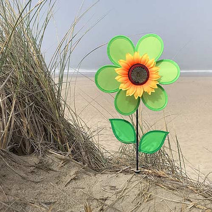 A decorative flower-shaped garden stake with green outer petals, a yellow and orange center, and two green leaves, planted in sand next to beach grass with a beach in the background.