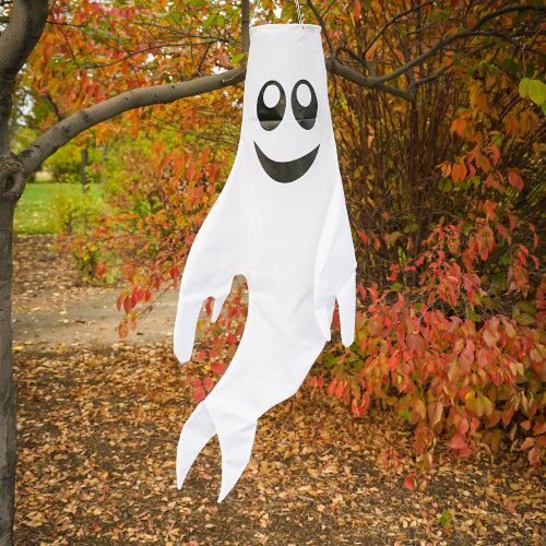 A white ghost-shaped hanging decoration with black eyes and a black smiley mouth, suspended from a tree branch outdoors in autumn foliage.