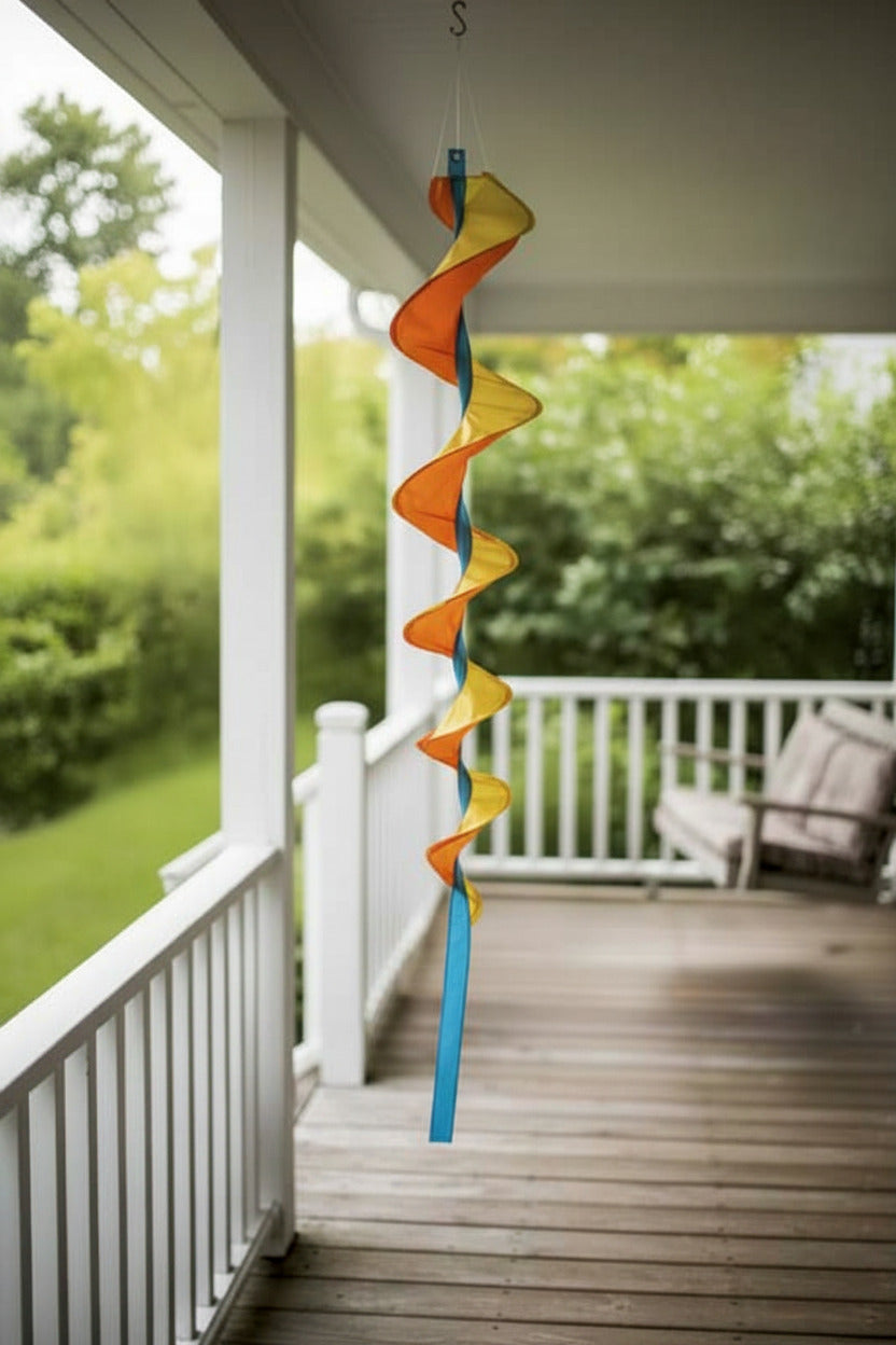 A spiral wind spinner with orange, yellow, and blue translucent panels hanging from a porch ceiling on a wooden deck with a white railing and green garden in the background.