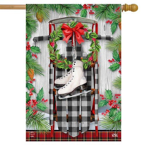 Decorative flag with ice skates hanging on a plaid patterned sled, surrounded by ivy and berries on a wooden background.